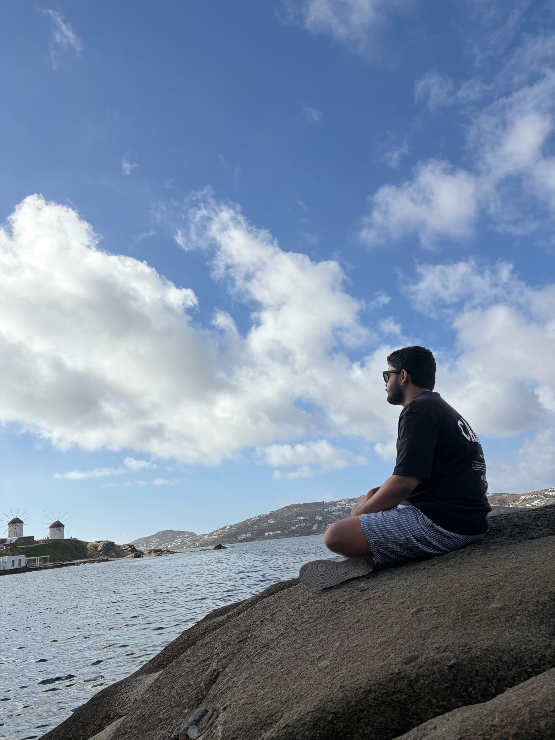 Yesith sitting alone on a rock by the sea in Mykonos, windmills in the distance